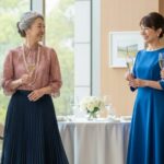 ホテルの窓際で笑顔でシャンパンを片手に会話を楽しむ60代の女性4人。それぞれがグリーン、ピンク、ブルー、ベージュの品のある同窓会ファッションを着こなしている。